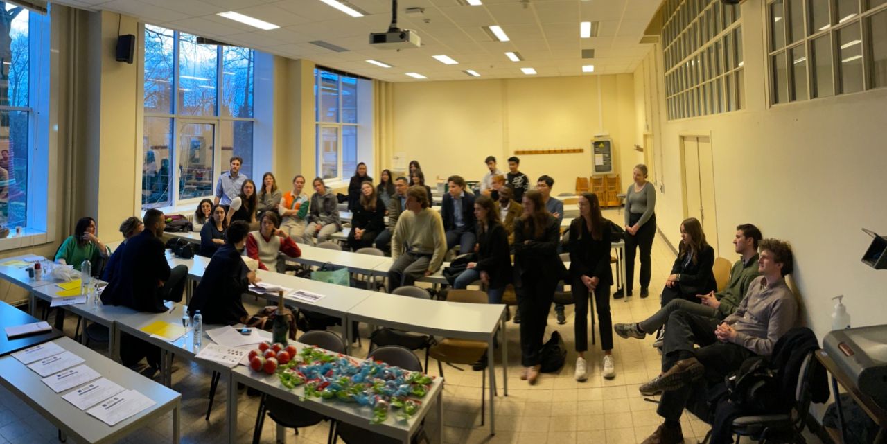 A group of people gathered in a classroom setting, sitting and standing around tables, engaged in a discussion.